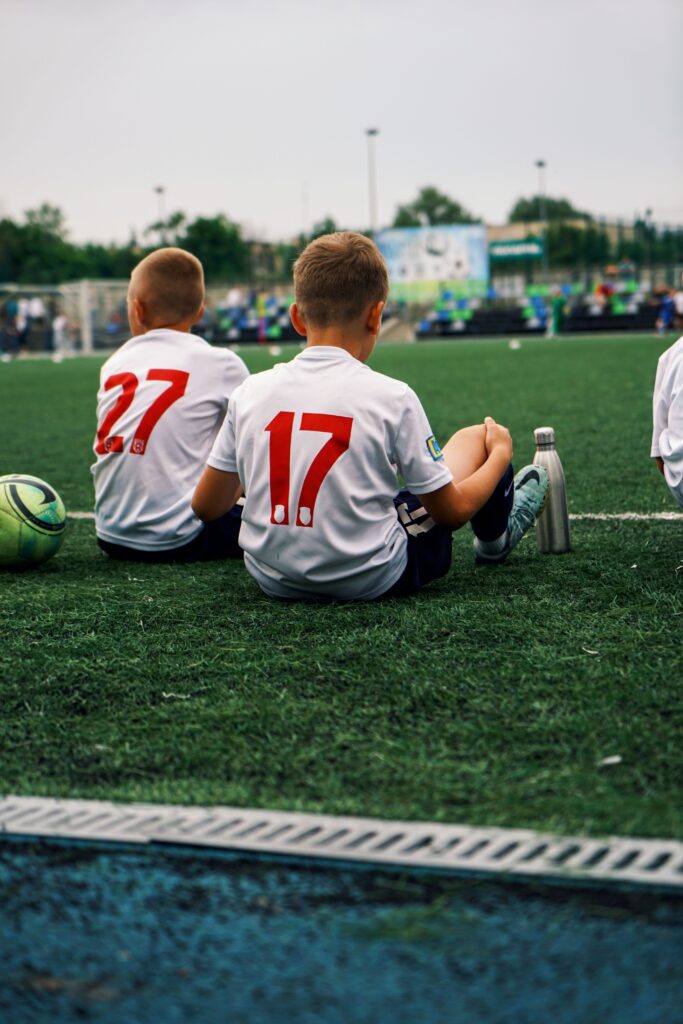 Boys in soccer jerseys sitting on a turf field during a summer game.